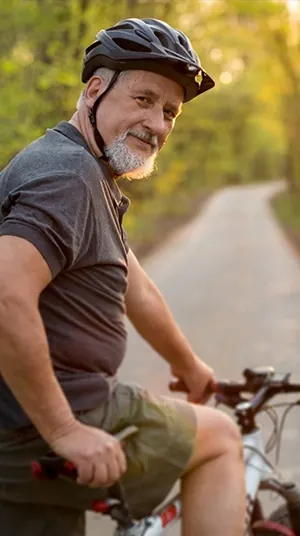 Man in his 60s in a bike helmet sitting on his bicycle on a wooded trail