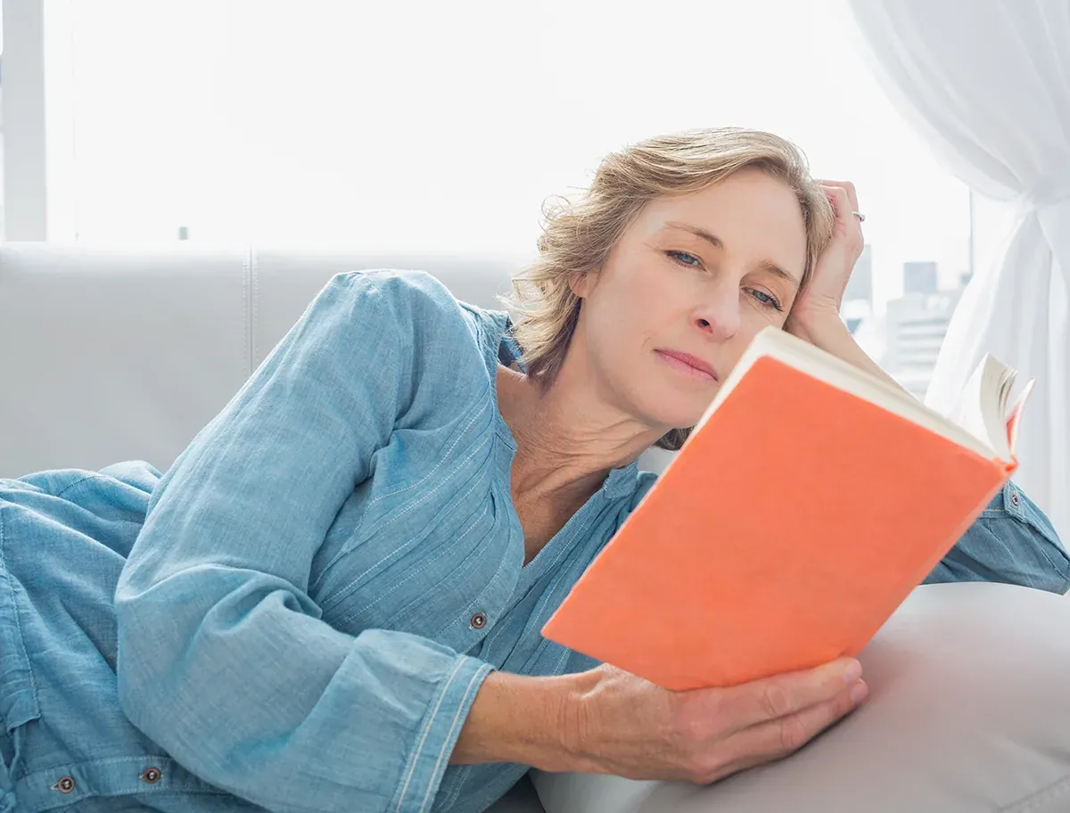 Woman in her 50s relaxing on a sofa, reading a book without glasses