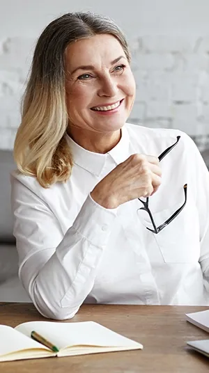 Smiling woman in her 50s at a desk, holding her reading glasses in one hand